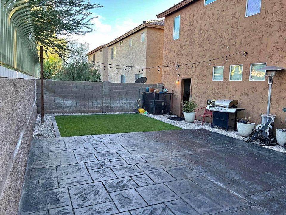 A backyard with a large slate patio, artificial grass, and a string of lights overhead. There are two-story homes with stucco walls surrounding the space. Against the house in Phoenix, there's a barbecue grill, potted plants, and various gardening supplies.