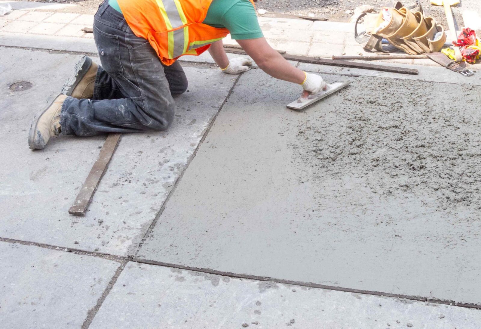 A construction worker in an orange vest and green shirt is kneeling on the ground, smoothing freshly poured concrete with a trowel on a sidewalk. Various tools and equipment are scattered nearby, showcasing the meticulous work of East Valley Concrete, your trusted concrete contractor in Chandler AZ.