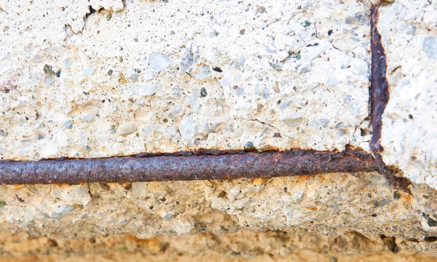 A close-up of deteriorating concrete with visible rusted rebar exposed. The rough, cracked surface reveals the wear and damage over time. The rusty metal bar runs horizontally through the middle of the image, a stark contrast to the quality work offered by decorative concrete contractors in Phoenix.