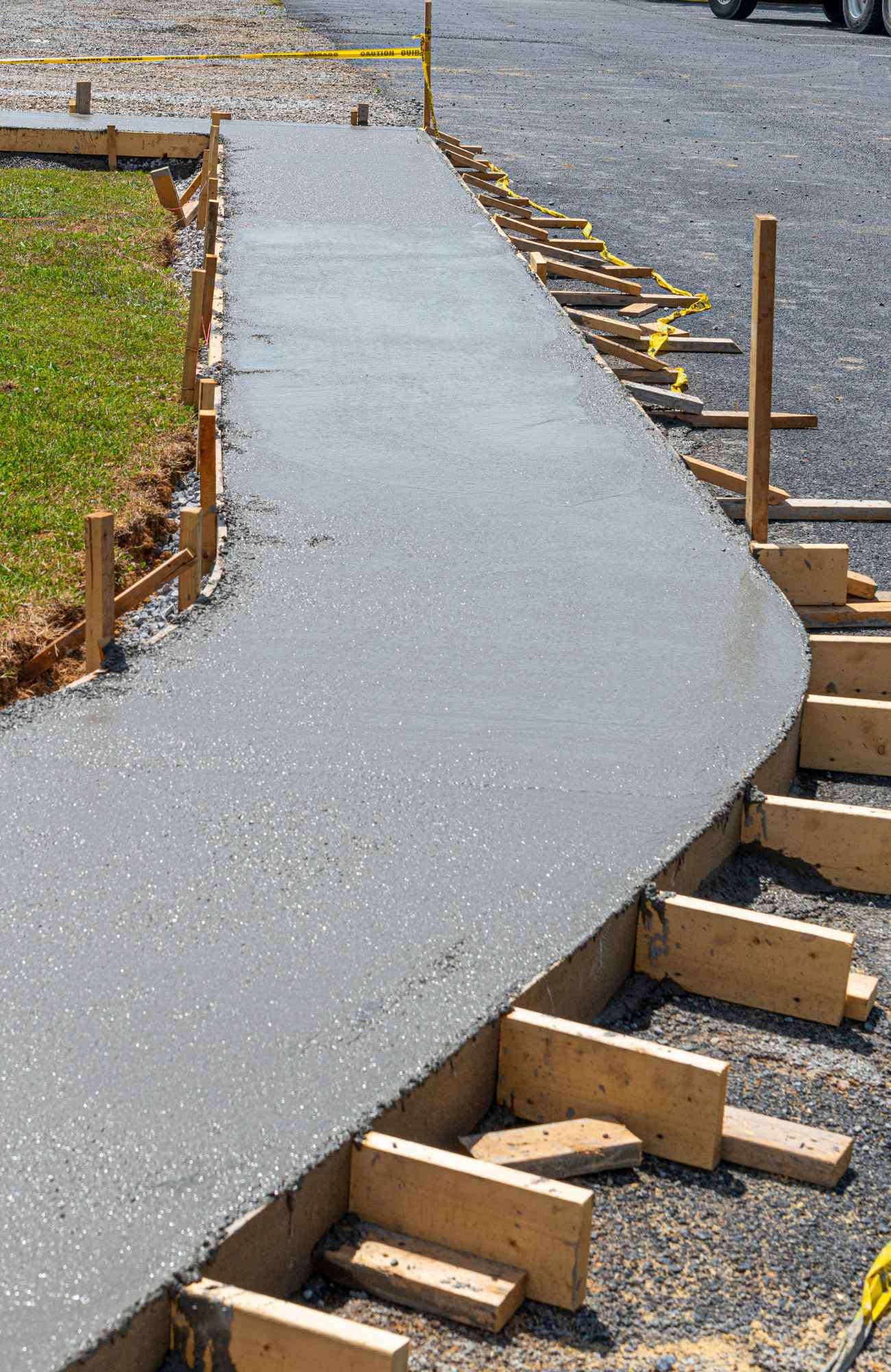 A newly poured curved concrete sidewalk in Paradise Valley is still wet, bordered by wooden forms to hold its shape. Yellow caution tape is visible in the background, and parts of the surrounding grassy and gravel areas are seen. The clear sky suggests a sunny day, expertly crafted by a licensed concrete contractor.