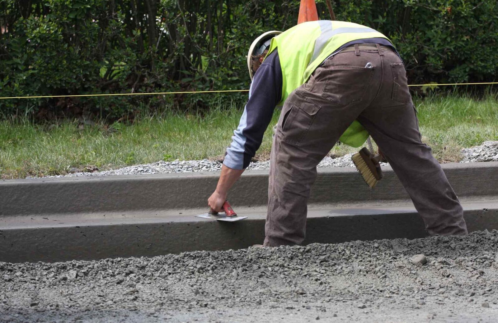 A construction worker wearing a reflective vest and helmet smooths wet concrete with a trowel while kneeling on a newly constructed curb. A brush is tucked into their back pocket, and greenery is visible in the background. Licensed concrete contractors in Phoenix offer free quotes for decorative concrete projects like these.