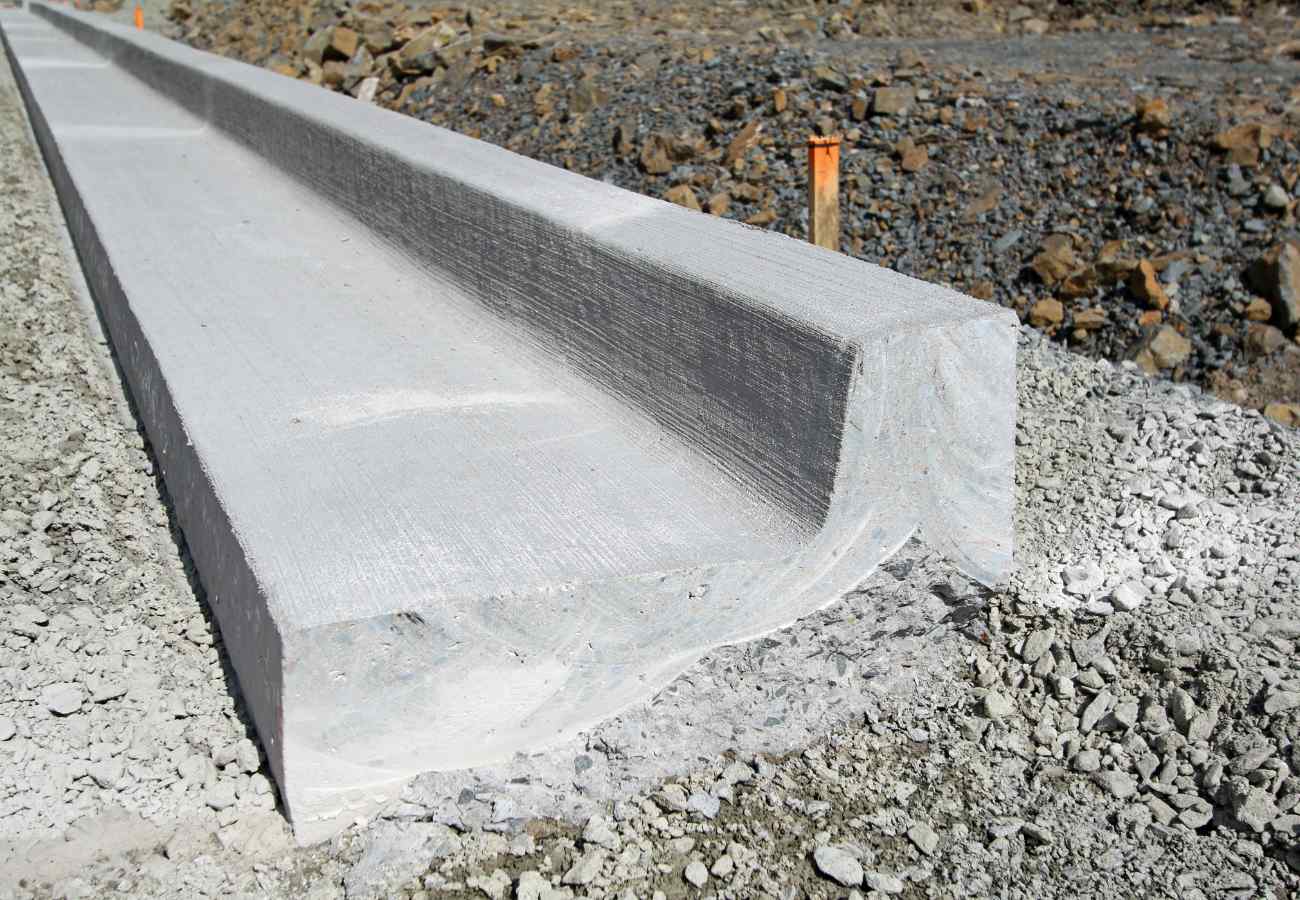 A close-up image of a concrete curb being installed in a construction area in Tempe, AZ. The curb has a clean, straight edge and is surrounded by gravel and earth. Orange markers are visible in the background, indicating the layout of the construction site. For all your concrete needs, contact us for free quotes.