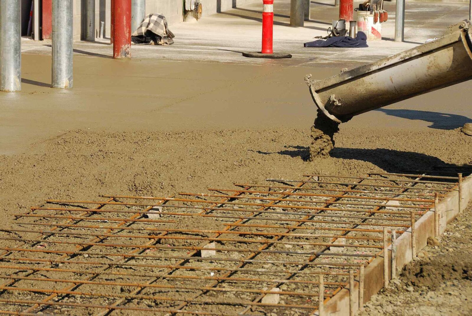 Concrete is being poured from a chute onto a metal rebar grid, forming a fresh slab. The surrounding construction area in Phoenix features scaffolding, pipes, and various materials. A worker's jacket and some tools are visible in the background. Decorative Concrete Contractors offer free quotes for similar projects.