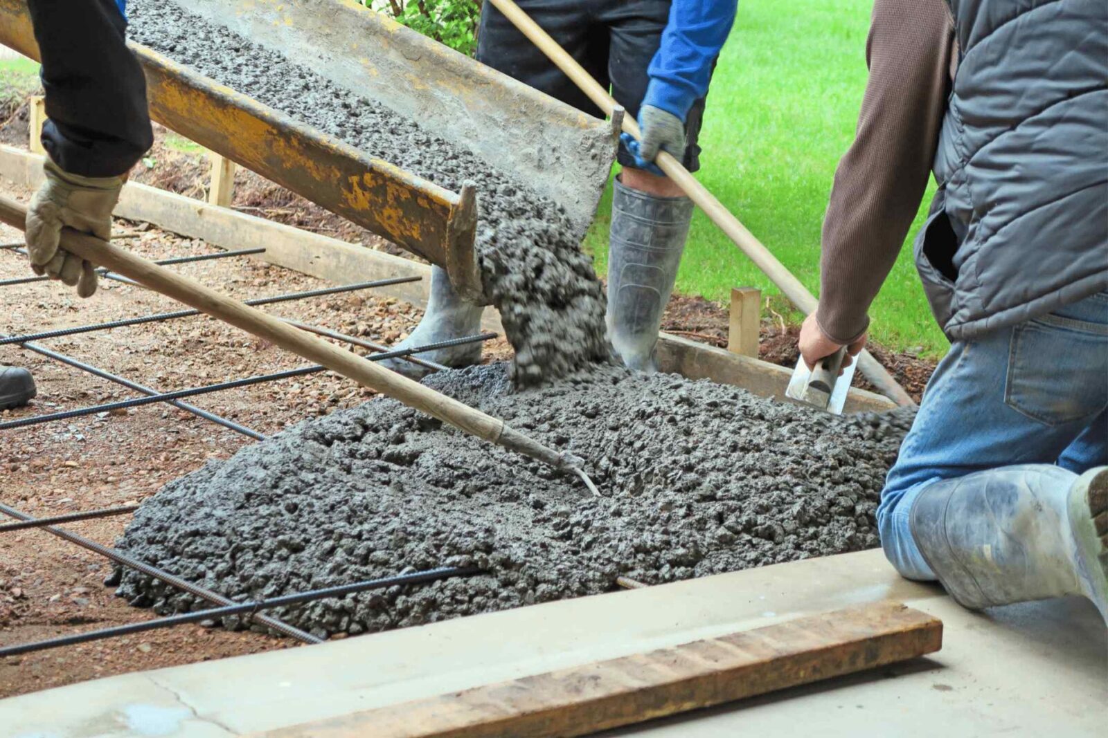 Three construction workers, part of a licensed concrete contractor crew, are using tools to spread and level wet concrete being poured from a chute onto a rebar-reinforced base. They are preparing the foundation for a new structure in Scottsdale. The scene takes place outdoors on a grassy yard.