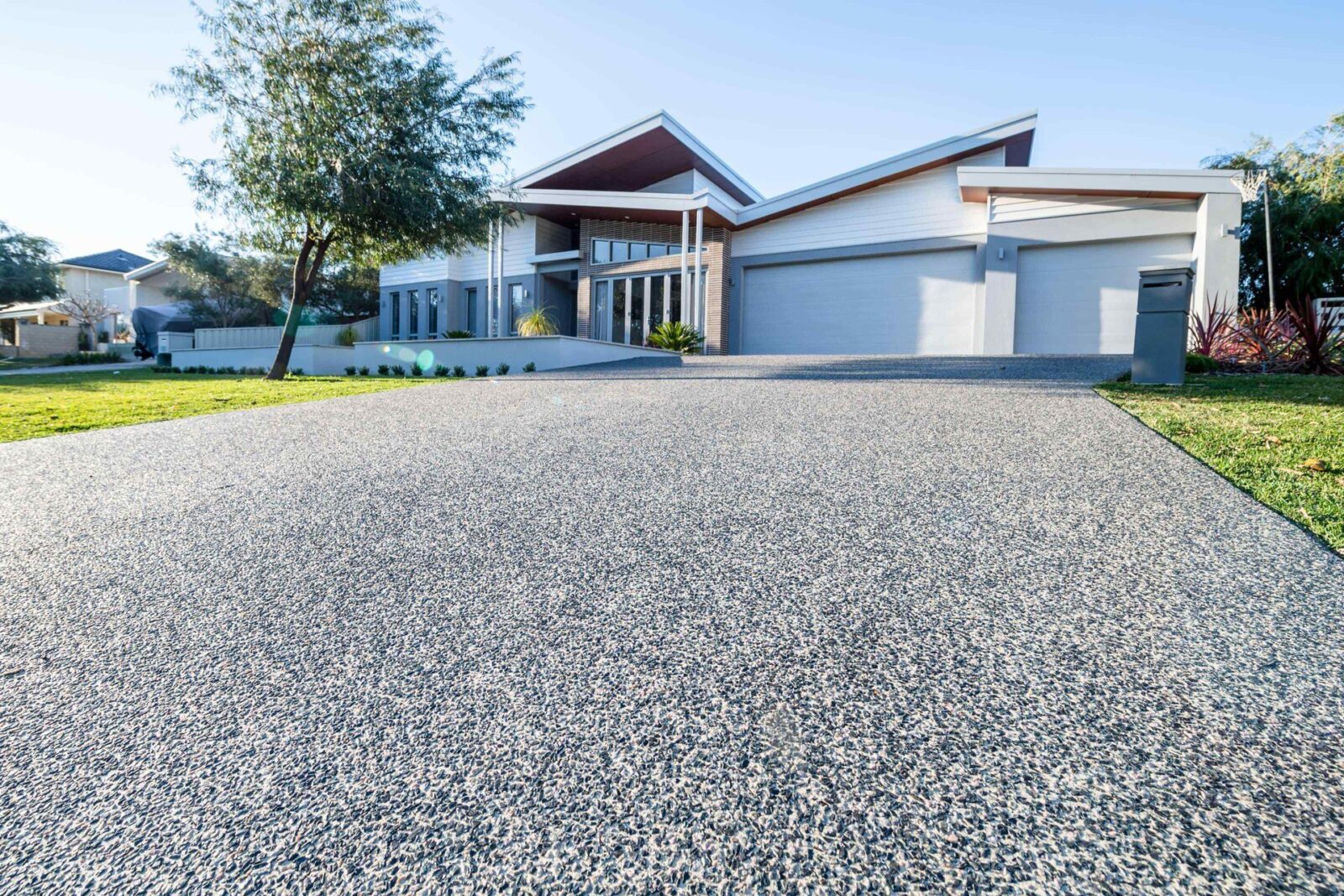 Modern house with clean architectural lines, a spacious concrete driveway by East Valley Concrete, neatly manicured front lawn, and large windows. The house features a flat and sloping roof design, a three-car garage, and is surrounded by minimalist landscaping with a few trees and shrubs. Free quotes available.