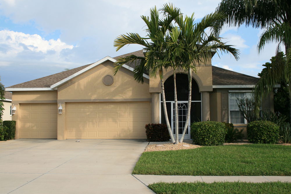 A single-story, tan stucco house in Tempe, AZ with a two-car garage, palm trees in the front yard, and a neatly manicured lawn. The house features a peaked roof, a covered entrance area, and a wide driveway extending towards the street for all your concrete needs.