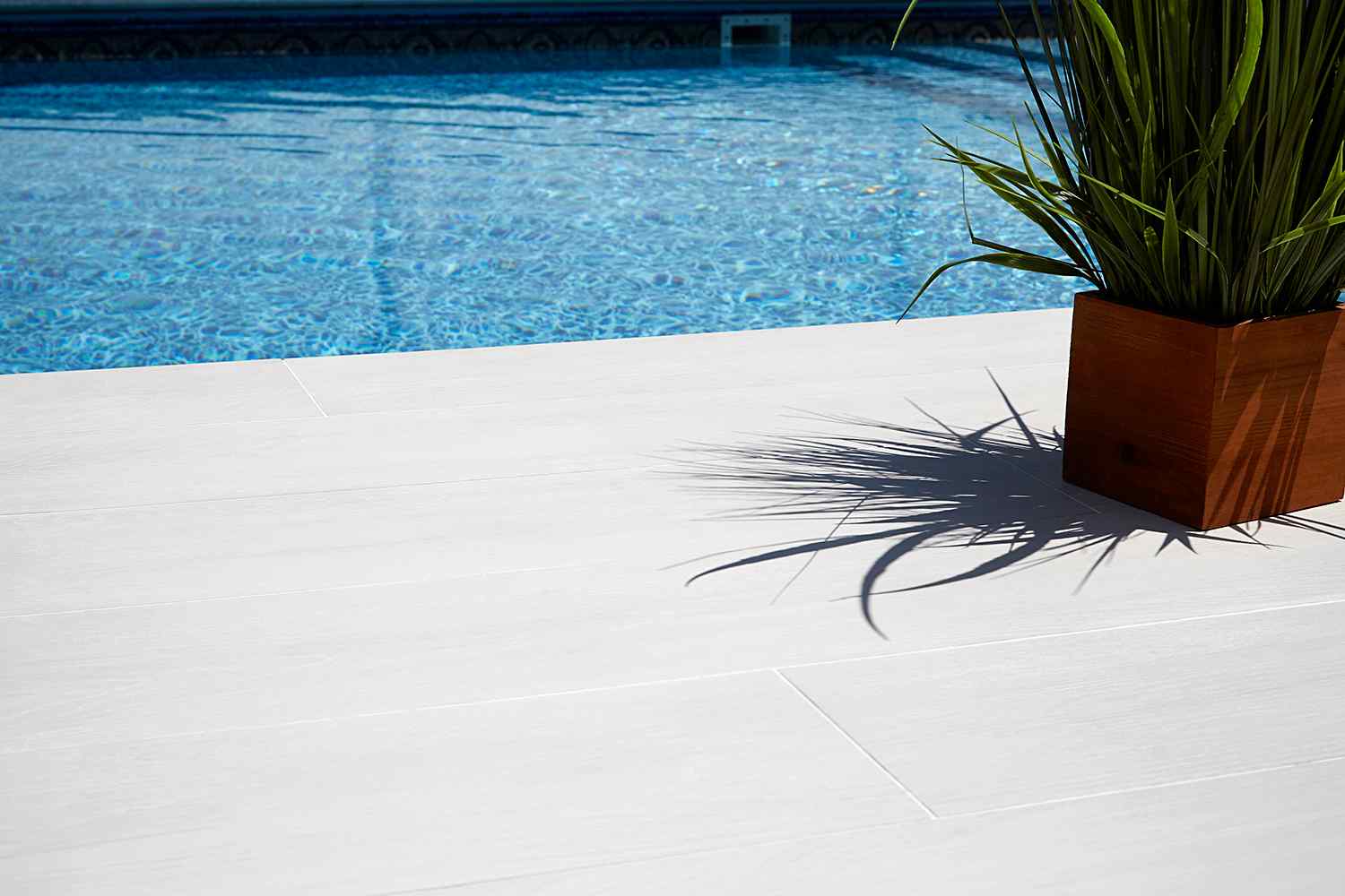 A close-up view of a swimming pool with clear blue water and a wooden deck, complemented by decorative concrete elements. A potted green plant in a wooden planter casts a shadow on the deck, creating a relaxing and inviting outdoor scene ideal for licensed, insured, and bonded projects.