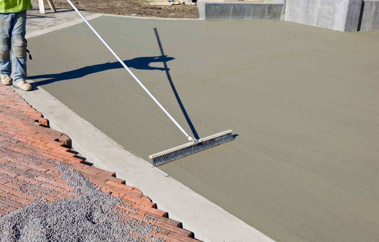 A worker in jeans, from East Valley Concrete, uses a long-handled tool to smooth and level freshly poured concrete bordering a brick pathway. The concrete is light gray and still wet, and there is a pile of gravel nearby. In the background, Chandler AZ's dry landscape and building materials can be seen.