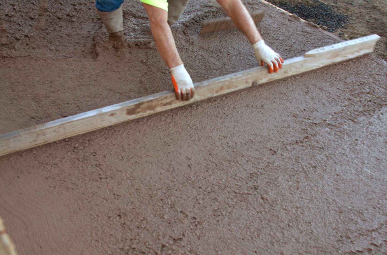 A licensed and insured construction worker in Mesa, AZ uses a long, flat tool to smooth a large area of wet concrete. Both hands grasp the tool's handles, and they wear gloves. The freshly poured concrete is damp and uneven, ensuring quality work by local concrete contractors.