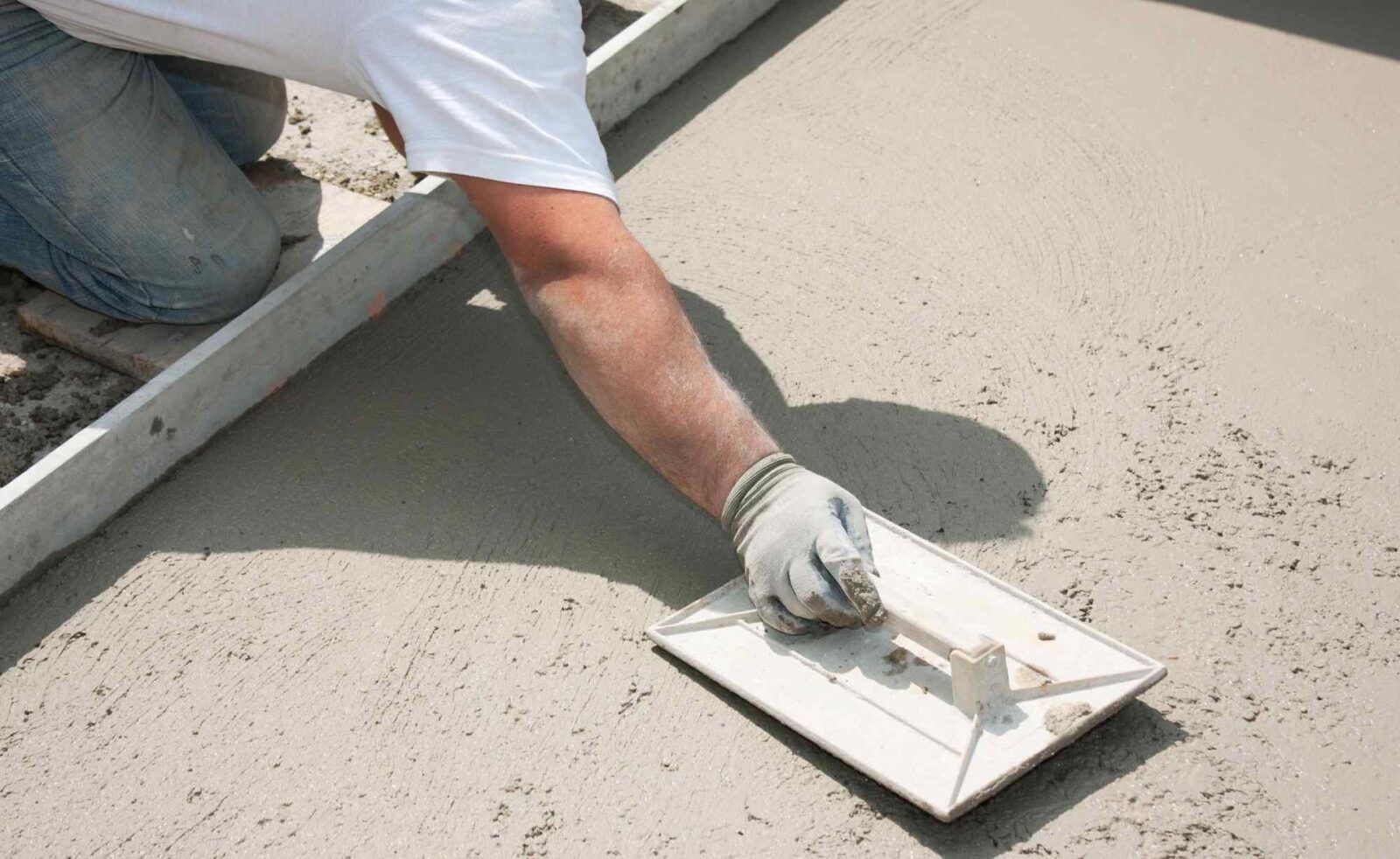 A person wearing a white shirt and gloves smooths wet concrete using a trowel tool. They are kneeling on the ground, and the concrete appears freshly poured. The scene looks like part of a larger construction project in Mesa AZ by experienced concrete contractors offering free quotes.