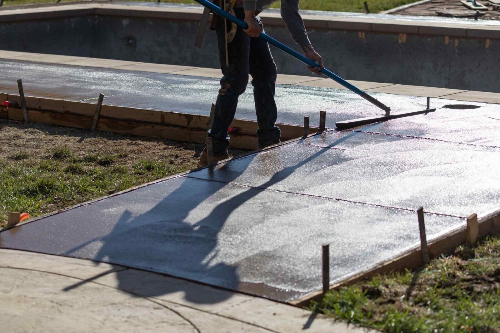 A licensed concrete contractor smooths out newly poured concrete on a pathway near an empty pool in Scottsdale using a long tool. The concrete is framed by wooden forms, and the surrounding area has grass and dirt. The sun casts shadows of the worker and the tool onto the wet concrete.