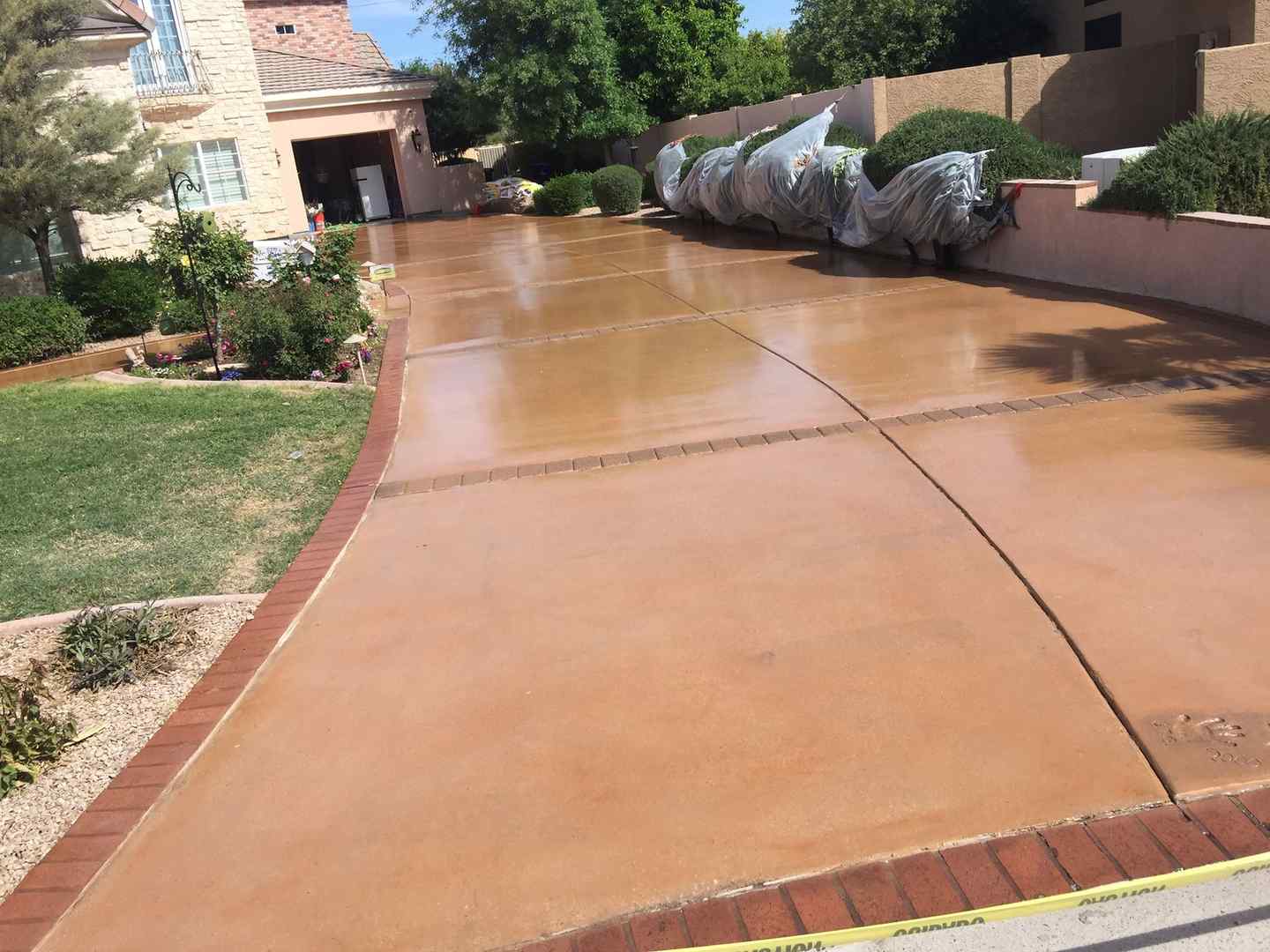 A clean, wide, tan concrete driveway with brick borders, expertly crafted by a local concrete contractor in Gilbert AZ, leads to a large garage in a residential area. The driveway is lined with green shrubs and covered items on the right. A light brown two-story house with manicured lawn is visible on the left side. Free quotes available upon request.