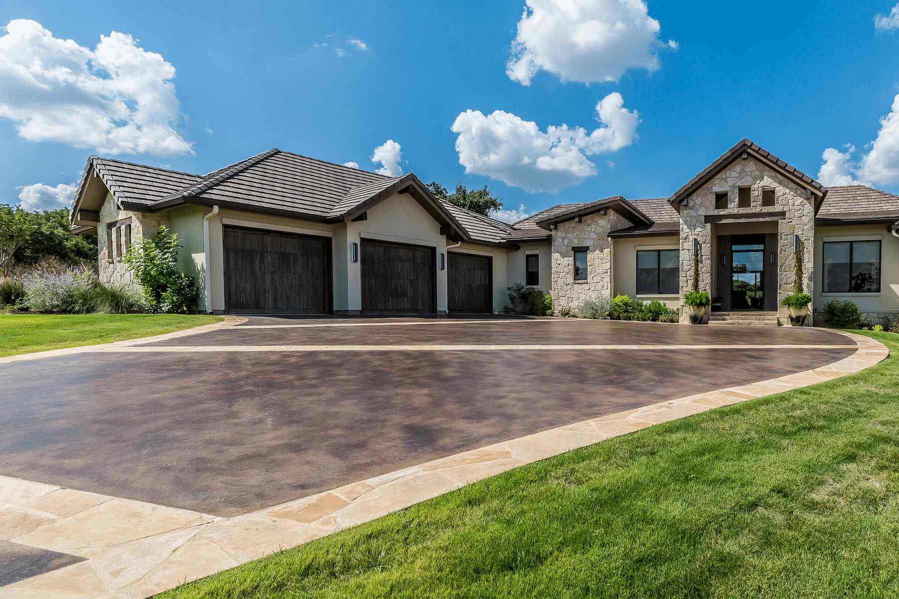 A large, modern single-story house with a stone and stucco exterior, dark wooden garage doors, and a wide, curved driveway crafted by a licensed concrete contractor. Nestled in Scottsdale, the property is surrounded by green grass and shrubbery under a bright blue sky with fluffy white clouds overhead.