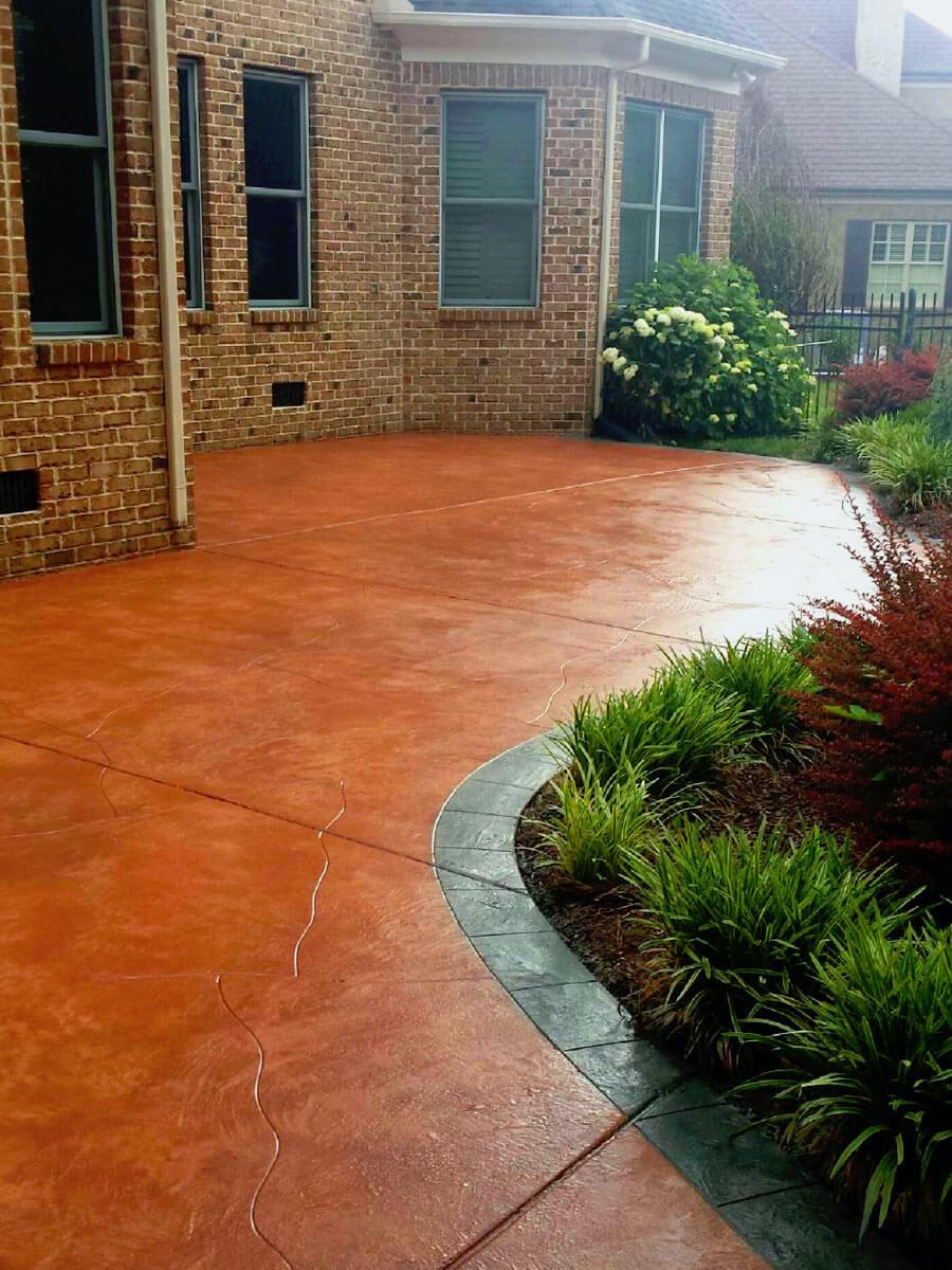 A backyard features a reddish-brown concrete patio bordered by a gray curved edge. The patio, masterfully crafted by decorative concrete contractors, is adjacent to a brick house with multiple windows. On the right, there's a lush garden with green shrubs and a blooming bush.