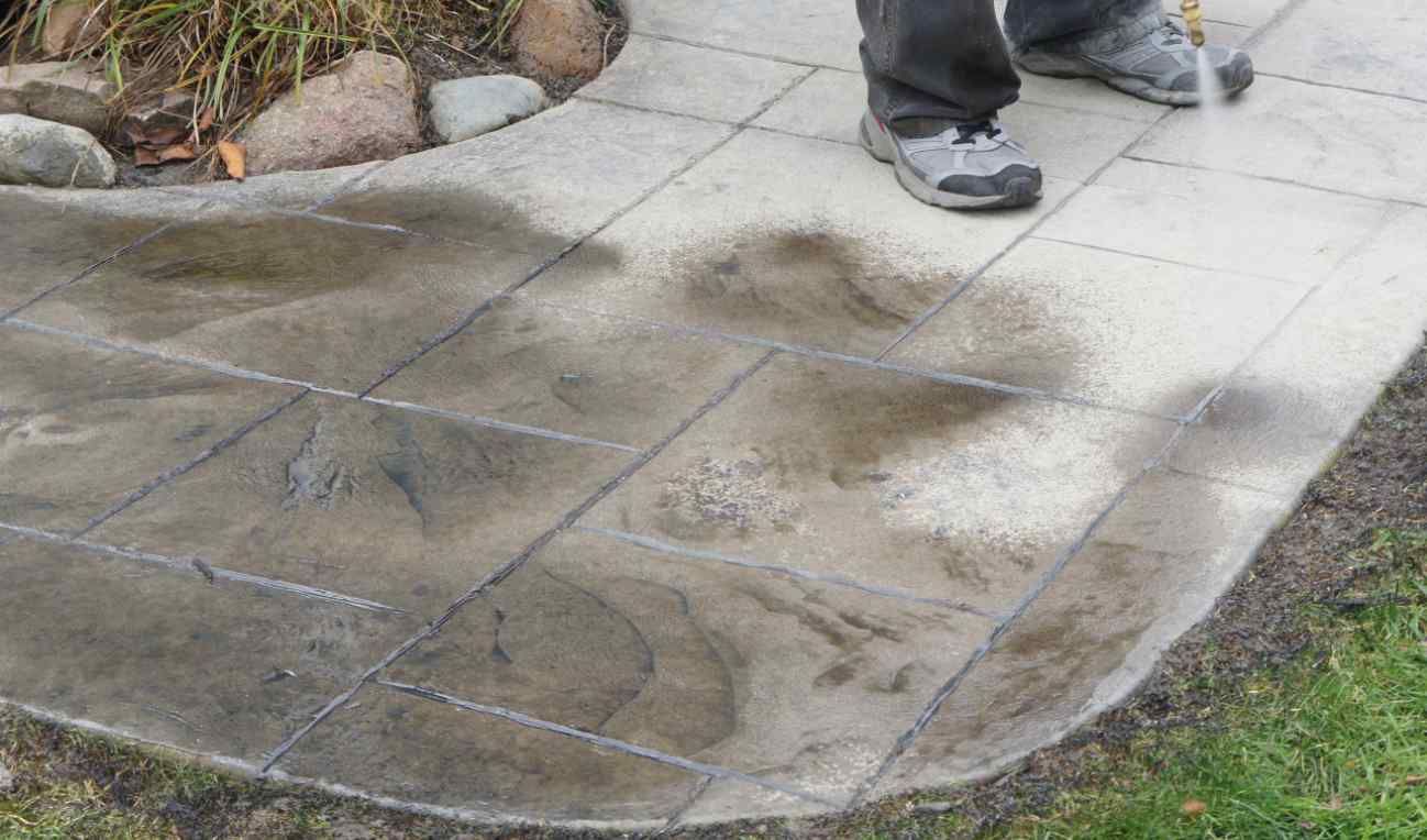 A person wearing grey sneakers and dark pants is power washing a tiled outdoor patio. The left side of the tiles appears dirty, while the right side is clean and wet from the power washing. Grass and rocks border the patio, making it a perfect project for Mesa AZ concrete contractors who offer free quotes.