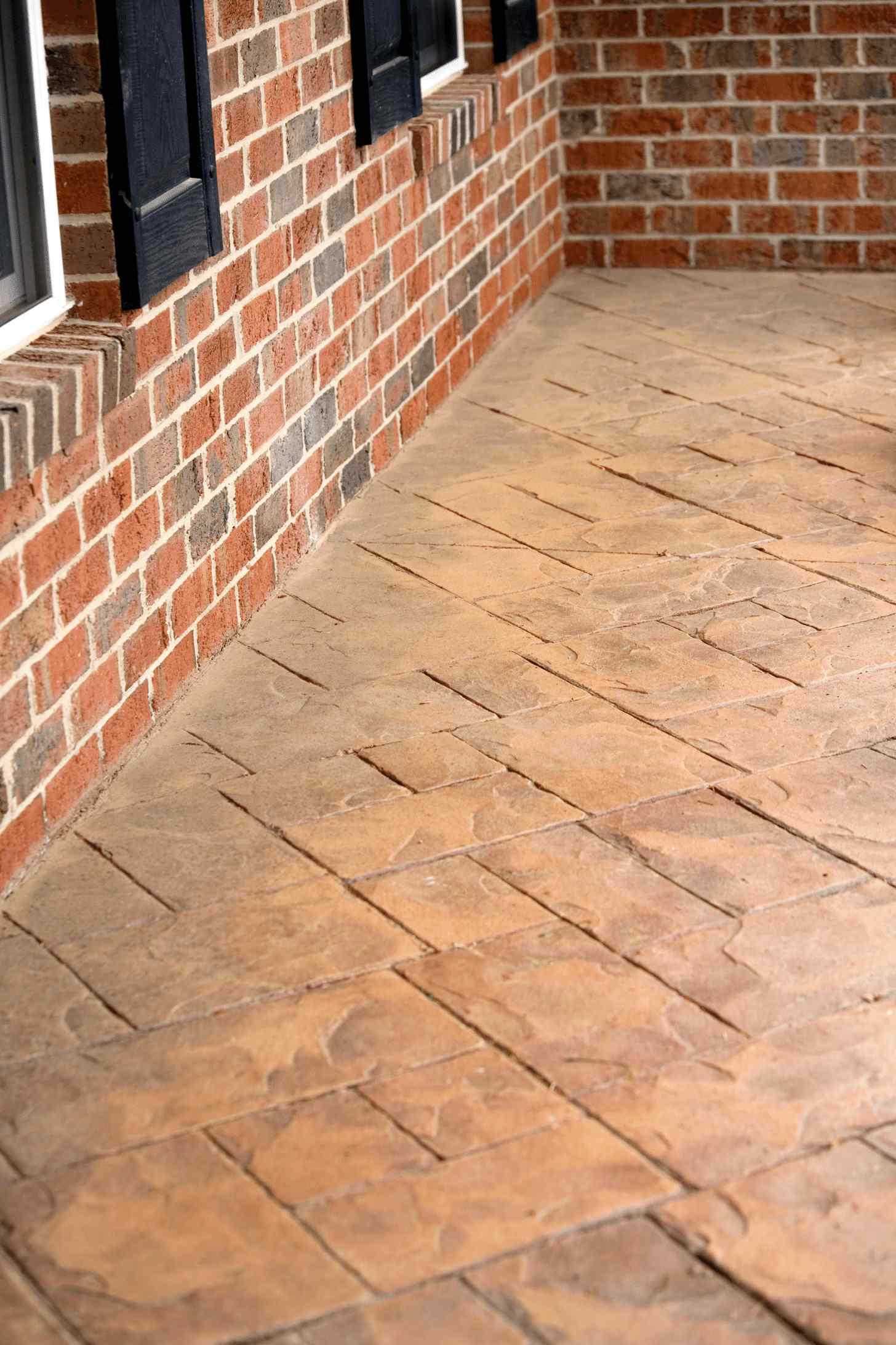 A close-up of a porch in Scottsdale featuring a textured, brick-patterned concrete floor next to a brick wall with windows. The concrete has a warm, brownish hue that complements the red and orange tones of the brick wall. Black shutters frame the windows. Contact our concrete contractor for free quotes today.