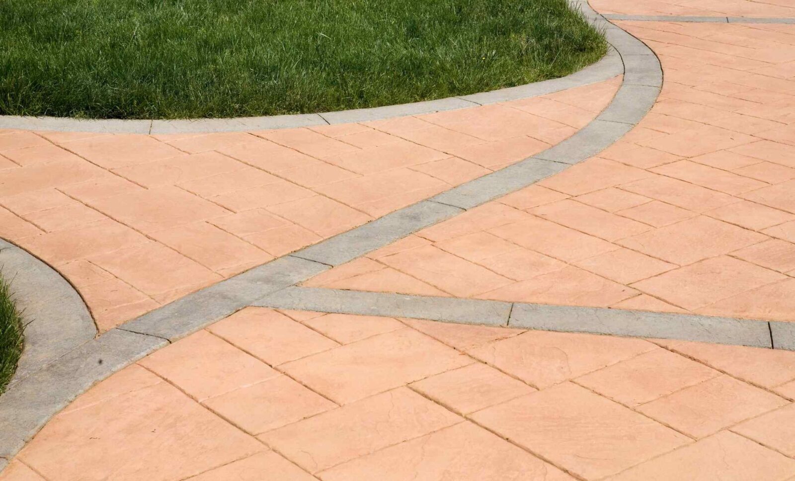 A patterned concrete walkway by East Valley Concrete features intersecting paths with a pinkish hue and gray borders, surrounded by vibrant green grass. The paths weave together, creating a visually appealing landscape design. For concrete in Tempe, AZ, contact us for free quotes.