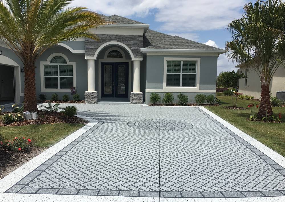 A spacious, modern house with a light gray exterior, featuring white trim, columns, and a dark double front door. The house has landscaped greenery and palm trees in the front yard, with a decorative patterned concrete driveway leading up to the entrance. Licensed and insured contractors provide free quotes for any renovations.