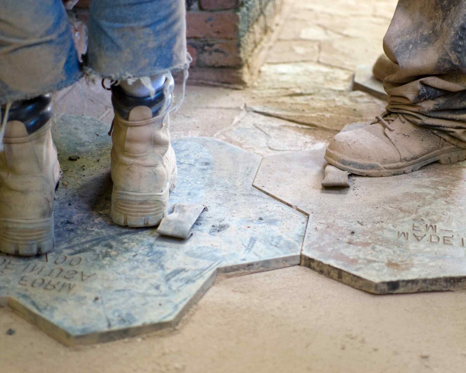 Close-up of two people wearing worn work boots standing on hexagonal stone tiles at a construction site in East Valley AZ. The image captures a portion of their legs and the dusty, unfinished floor with some scattered tiles and building materials, showcasing stamped concrete patio installation services.