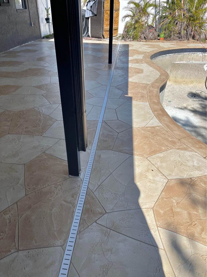 A partially tiled patio with beige and light brown hexagon and irregular-shaped tiles is shown. There is a linear drainage channel along the patio, and a partially visible, empty, plastered pool on the right. Potted plants are visible in the background. Decorative concrete contractors in Phoenix offer free quotes for such projects.