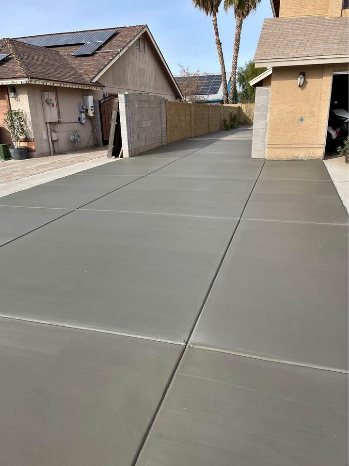 A freshly-paved concrete driveway leads to a garage of a beige house. Solar panels are visible on the roof of the neighboring home. The driveway is separated from the adjacent property by a cinder block wall, showcasing the work of licensed and insured decorative concrete contractors. Palm trees are seen in the background.