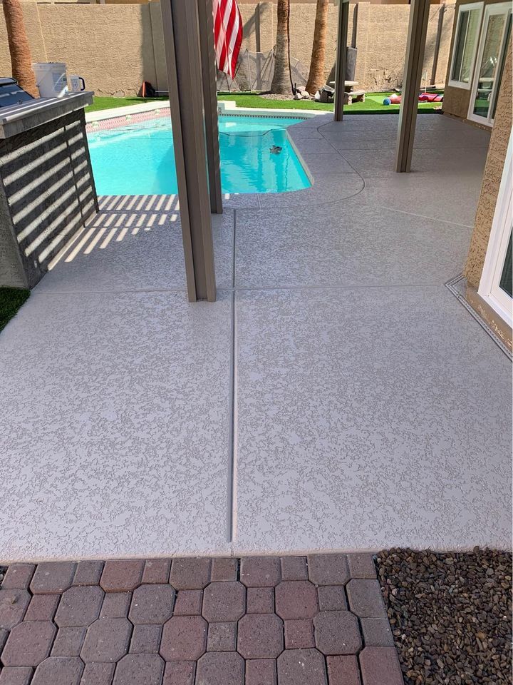 A backyard in Gilbert, AZ, featuring a swimming pool with clear blue water. Surrounding the pool is a textured concrete patio designed by East Valley Concrete, with a pergola providing partial shade. Paver stones and artificial grass are alongside the patio, and an American flag hangs in the background.