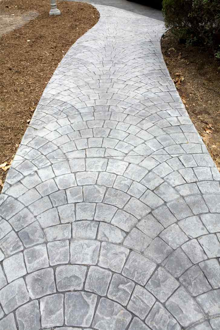 A curved, paved pathway made of durable interlocking stone tiles leads through a garden area. The stones are arranged in a fan-like pattern, creating a visually appealing design. The surrounding garden area is covered with mulch and has neatly trimmed shrubs. Contact us for free quotes on your next project.