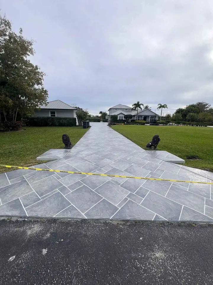 A paver installation transforms the driveway into an airplane runway, complete with yellow caution tape and two ornamental black sculptures. Framed by green lawns, the scene leads to a white house under a cloudy sky, merging creativity with functionality.