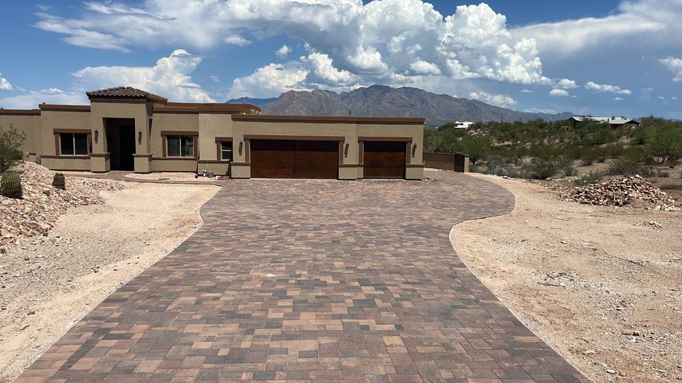 A modern, single-story house with a long brick driveway showcases expert paver installation amidst the desert landscape. The stucco exterior complements a three-car garage, while mountains grace the background under a partly cloudy sky.