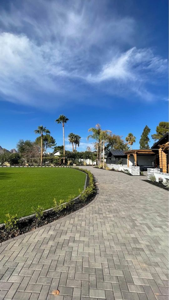 A paved pathway meanders through a lush garden with green grass and palm trees under a clear blue sky. To the right, a building with wooden accents complements adjacent patios. The scene is peaceful and vibrant with natural beauty.