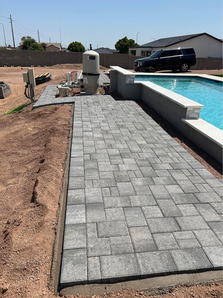 A newly constructed backyard showcases a swimming pool with expertly installed gray paver stones surrounding it. To the left, pool equipment is neatly arranged. In the background, a black SUV is parked near a low wall, completing this elegant outdoor retreat.