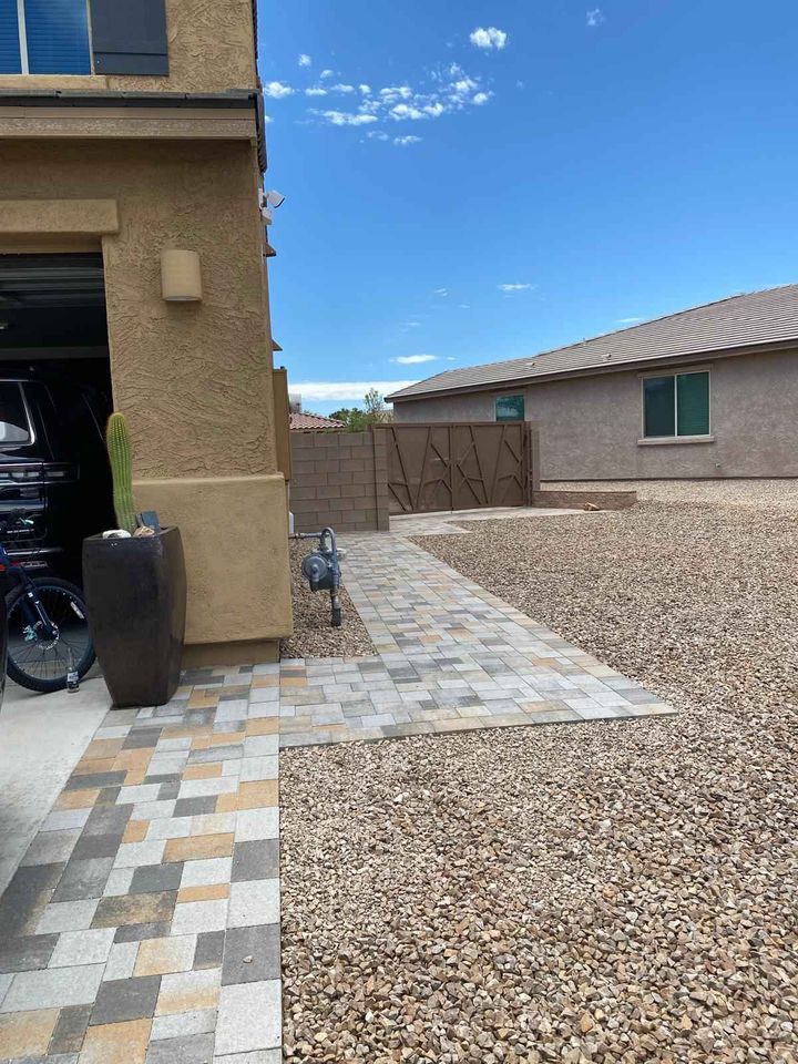 A house exterior features a partially open garage door, with a paved pathway leading through a gravel courtyard. A tall planter with a cactus sits near the garage, showcasing expert paver installation. The sky is clear with a few clouds.